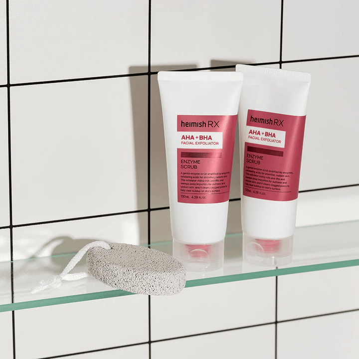 Dve tubi Heimish RX AHA + BHA Facial Exfoliator postavljeni na stekleno polico v kopalnici z belo ploščato steno. Poleg izdelkov leži plovec z belo vrvico. Elegantna in minimalistična slika nege kože.