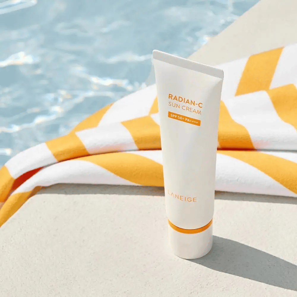 LANEIGE Radian-C Sun Cream stoji ob robu bazena z rumeno-belim črtastim brisačo v ozadju.