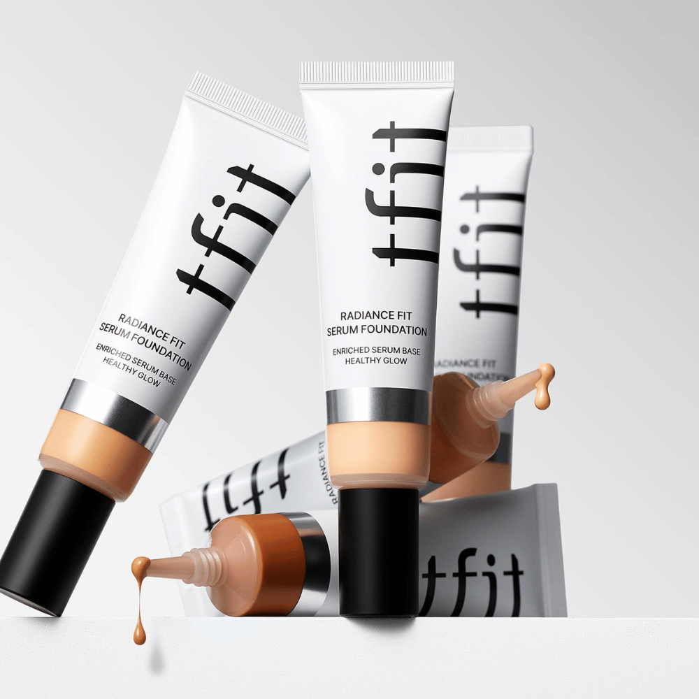 Več tub TFIT Radiance Fit Serum Foundation v različnih odtenkih, ena izmed njih z osnovo, ki kaplja, na svetlo sivi podlagi.