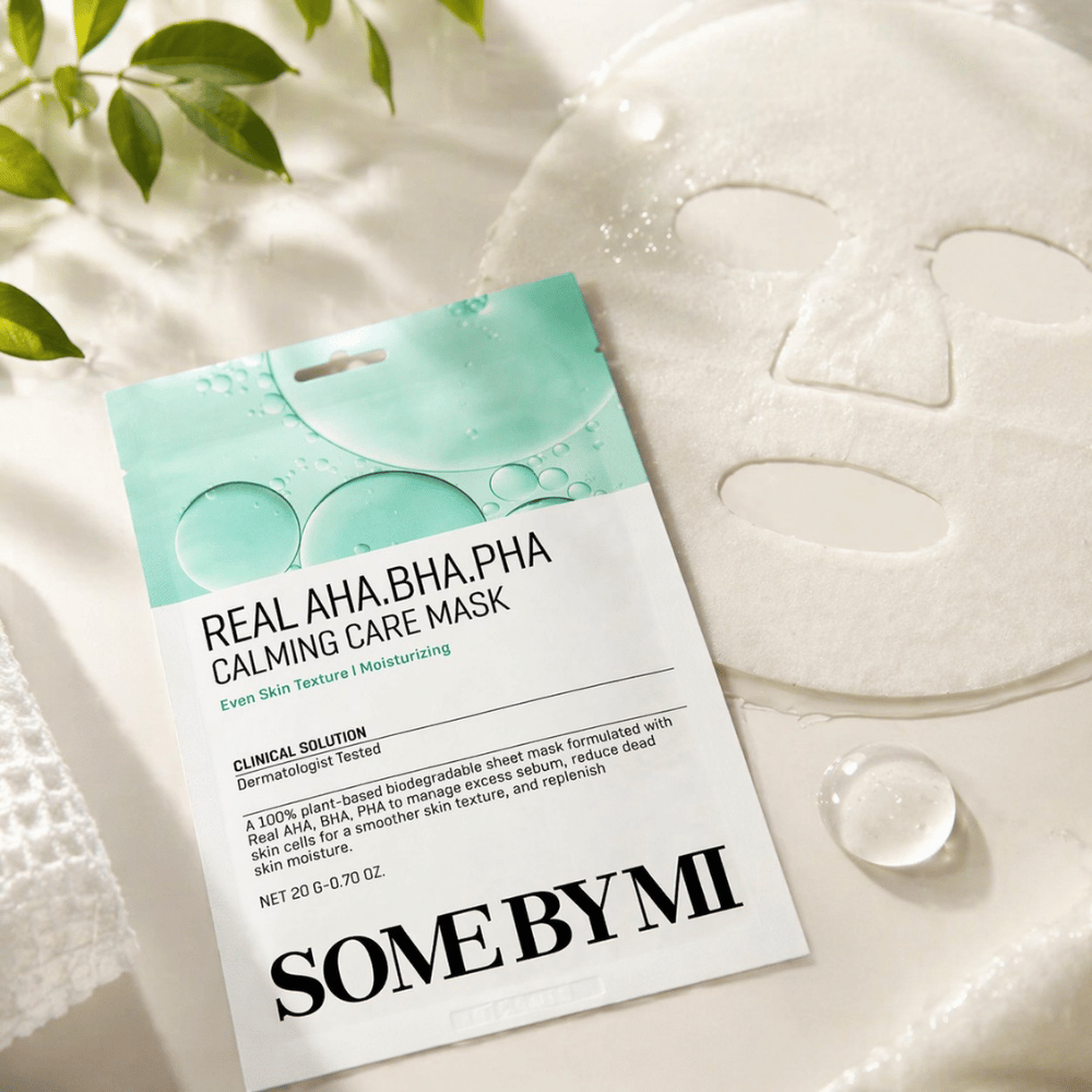 SOME BY MI Real AHA.BHA.PHA Calming Care Mask je prikazana skupaj z razvito listno masko. Okoli izdelka so vidne kapljice vlage in zeleni listi, ki poudarjajo občutek vlaženja, pomirjujoče nege in nežnega luščenja. Slika izpostavlja osredotočenost maske na enakomernejšo teksturo kože in izboljšano ravnovesje vlage.