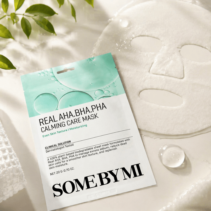 SOME BY MI Real AHA.BHA.PHA Calming Care Mask je prikazana skupaj z razvito listno masko. Okoli izdelka so vidne kapljice vlage in zeleni listi, ki poudarjajo občutek vlaženja, pomirjujoče nege in nežnega luščenja. Slika izpostavlja osredotočenost maske na enakomernejšo teksturo kože in izboljšano ravnovesje vlage.