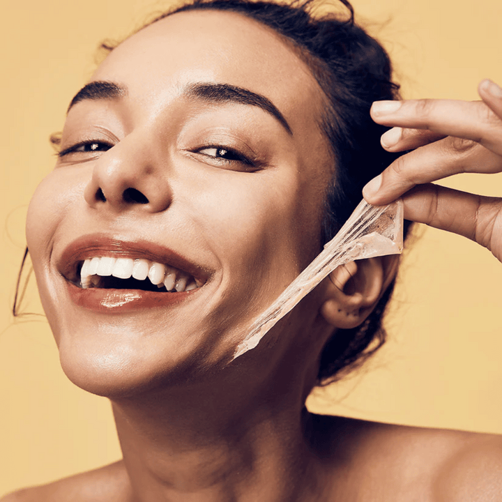 Slika prikazuje žensko z velikim nasmehom, ki si odstranjuje peel-off masko z obraza. Ženska izgleda vesela in zadovoljna, medtem ko uporablja lepotni izdelek, kar prenaša občutek dobrega počutja in nege kože.