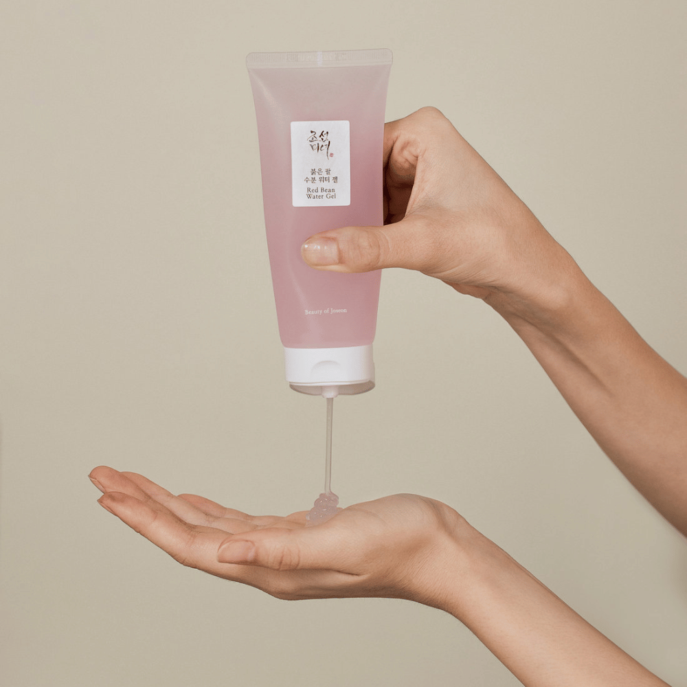 Slika prikazuje osebo, ki drži tubo "Red Bean Water Gel" iz Beauty of Joseon in iztiska prozoren gel v svojo roko. Izdelek je videti namenjen za nego kože in ima privlačno, nežno rožnato embalažo.