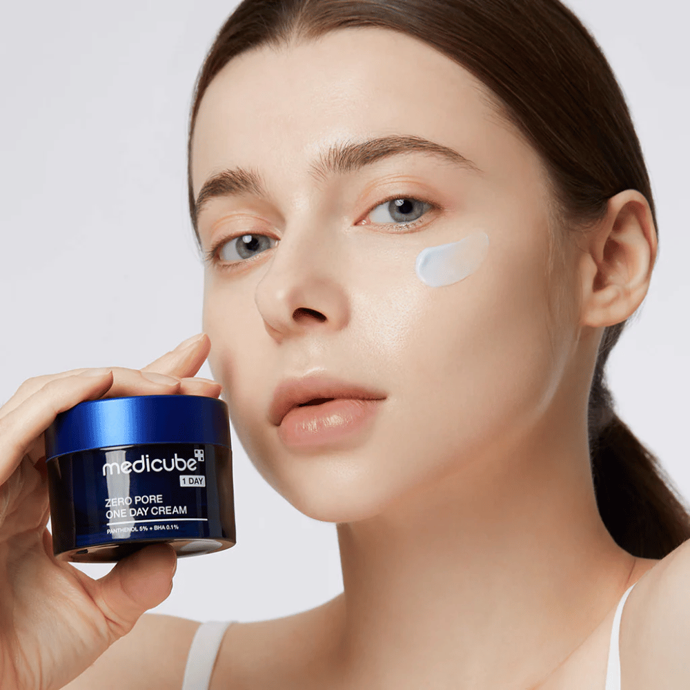 Ženska z naravnim videzom drži Medicube Zero Pore One Day Cream. Majhna količina kreme je nanesena na njenem licu. Izdelek je zasnovan za izboljšanje teksture kože in zmanjšanje velikosti por, z lahko in vlažilno formulo.
