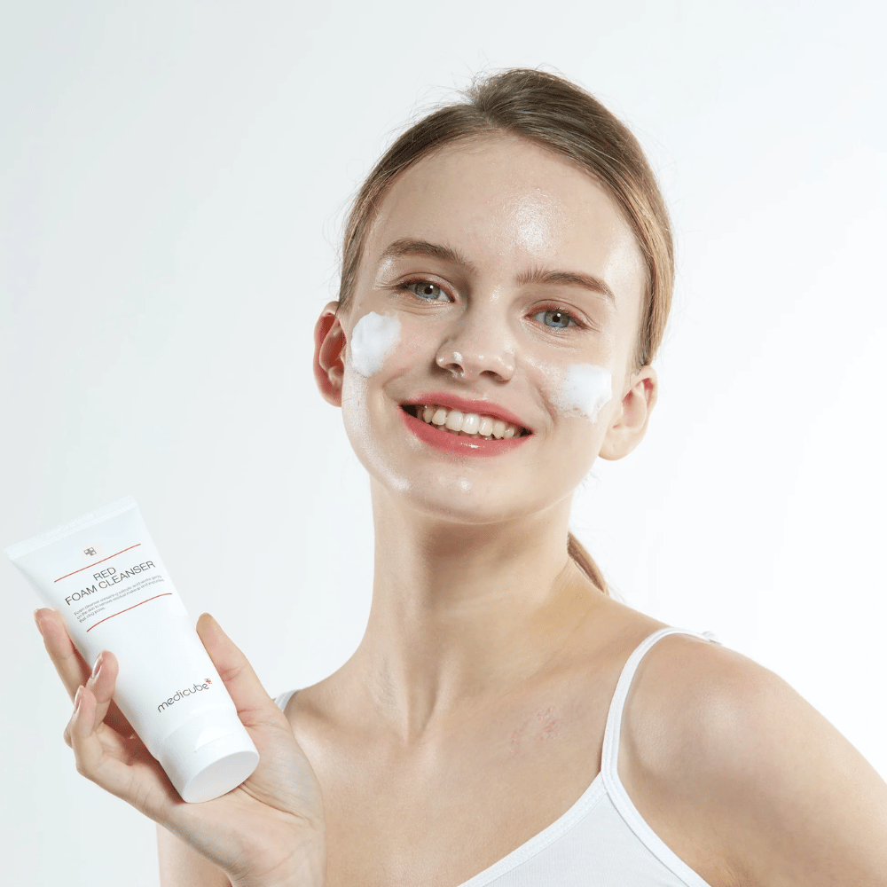 Mlada ženska s čistilno peno na obrazu drži tubo Medicube Red Foam Cleanser in se nasmehne kameri. Oblečena je v belo majico brez rokavov in ozadje je svetlo in enostavno.