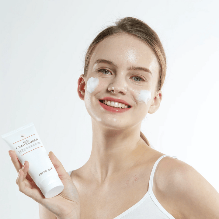 Mlada ženska s čistilno peno na obrazu drži tubo Medicube Red Foam Cleanser in se nasmehne kameri. Oblečena je v belo majico brez rokavov in ozadje je svetlo in enostavno.