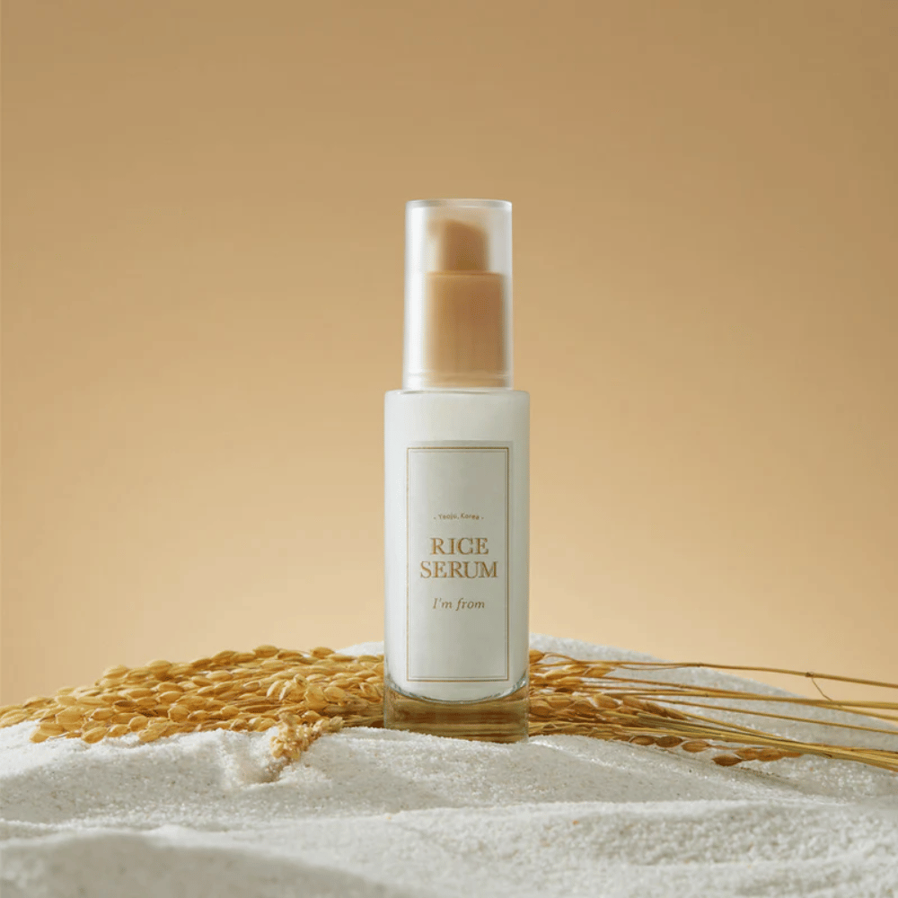 Steklenička z "I'm from" Rice Serum, obdana z riževimi zrni in riževimi klasi proti toplemu ozadju.