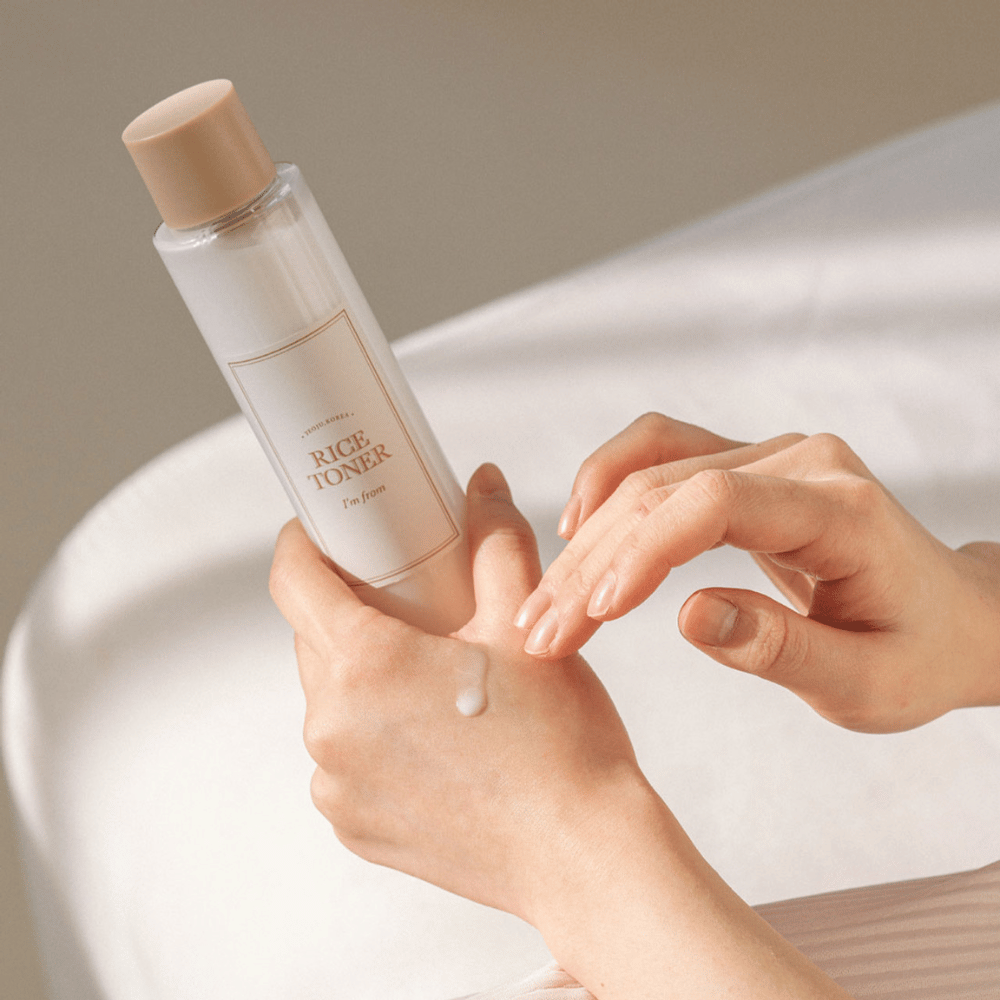 Roke držijo steklenico riževega tonerja z oznako "I'm from". Kapljica izdelka je nanesena na eno roko, medtem ko se druga roka pripravlja na nanos. Prizor je mehko osvetljen s toplo barvo.