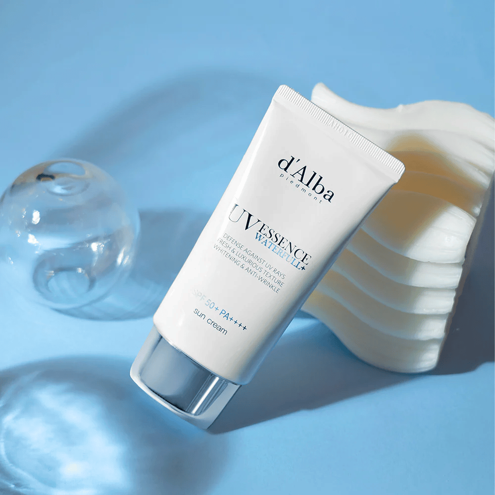 d'Alba UV Essence Waterfull+ Sun Cream z SPF 50+ PA++++ leži na modri podlagi z odsevi svetlobe in dekorativnimi elementi – poudarja sveže in vlažilne lastnosti kreme za sončenje.