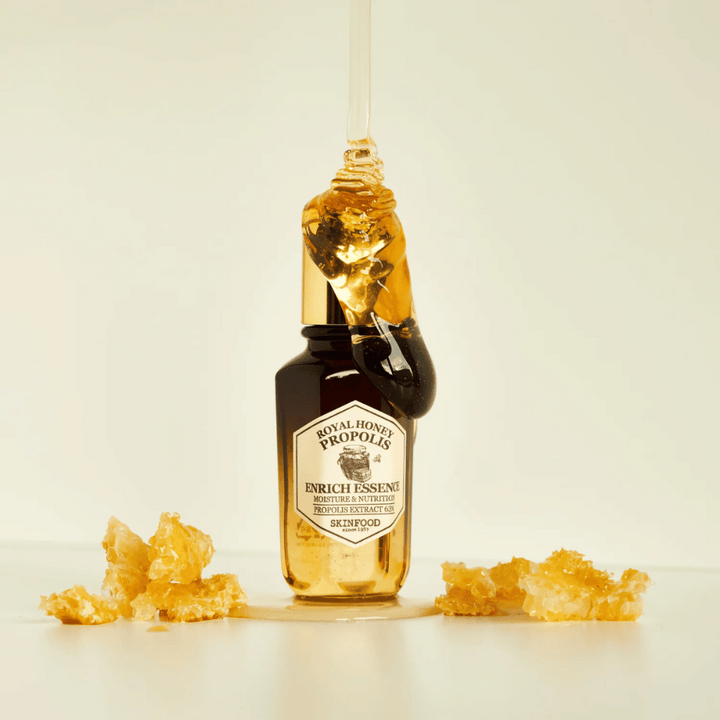 Slika steklenice iz stekla z Royal Honey Propolis Enrich Essence od Skinfood. Velika kapljica zlate medu teče po zlatem pokrovu steklenice. Okoli steklenice so majhni koščki satovja razpršeni po svetli površini. Esenca se oglašuje s poudarkom na vlagi in prehrani, s 63% izvlečkom propolisa.