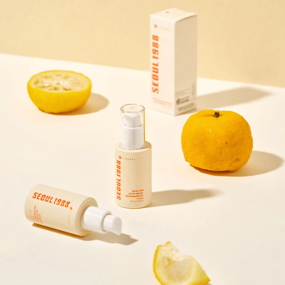 Več steklenic in embalaž Seoul 1988 Glow Serum, razporejenih s celimi in prepolovljenimi yuja sadeži v ozadju.