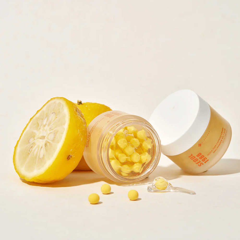 Odprta posoda z rumenimi kapsulami poleg razpolovljenega yuja-sadja, ki poudarja formulo bogato z vitaminom C.