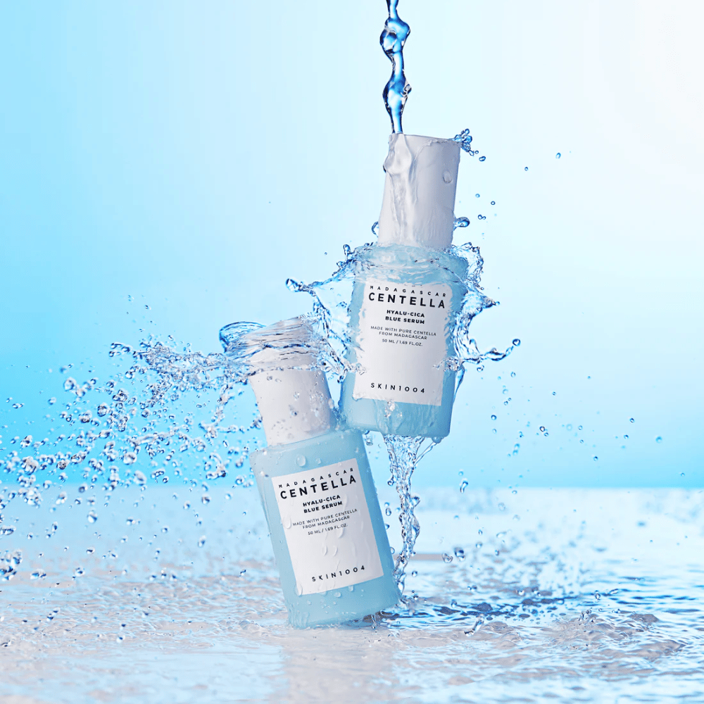 Dve steklenički SKIN1004 Madagascar Centella Hyalu-Cica Blue Serum sta zadeti z vodnim curkom na svetlo modrem ozadju.