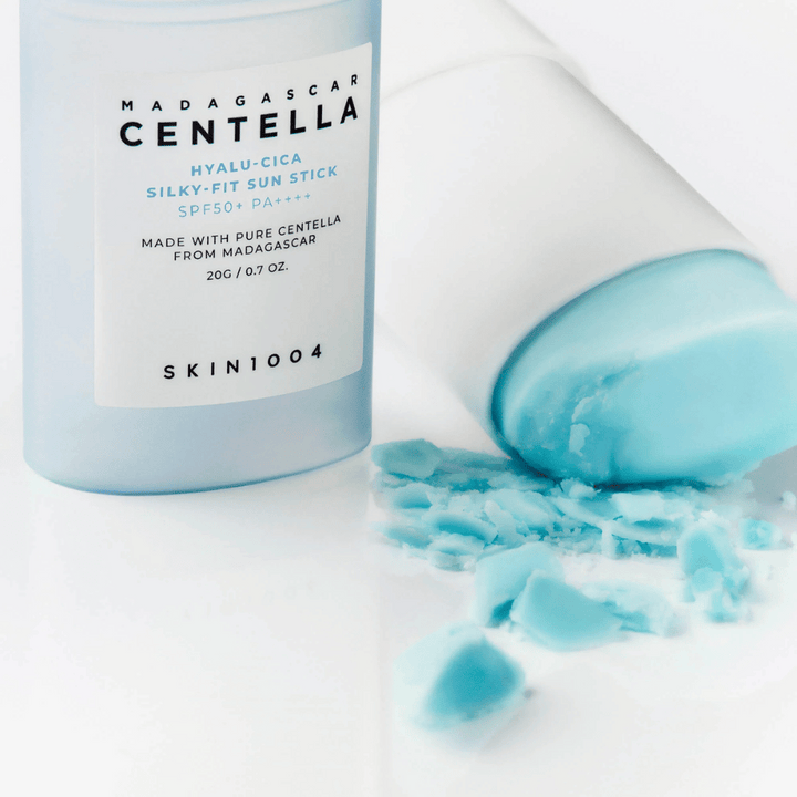 Zaščitni stik z SPF50+ leži odprt poleg svojega pokrova, vsebina je delno razmazana po beli površini. Besedilo na embalaži omenja sestavine iz Madagaskarja.