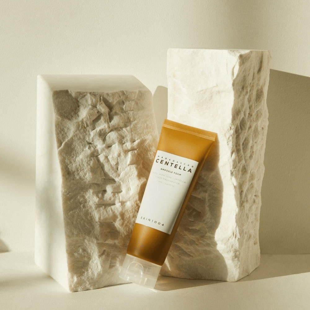SKIN1004 Madagascar Centella Ampoule Foam stoji ob svetlih kamnitih blokih v naravni sončni svetlobi, s svojo rjavo-belo zasnovo tube in elegantno minimalizmom.