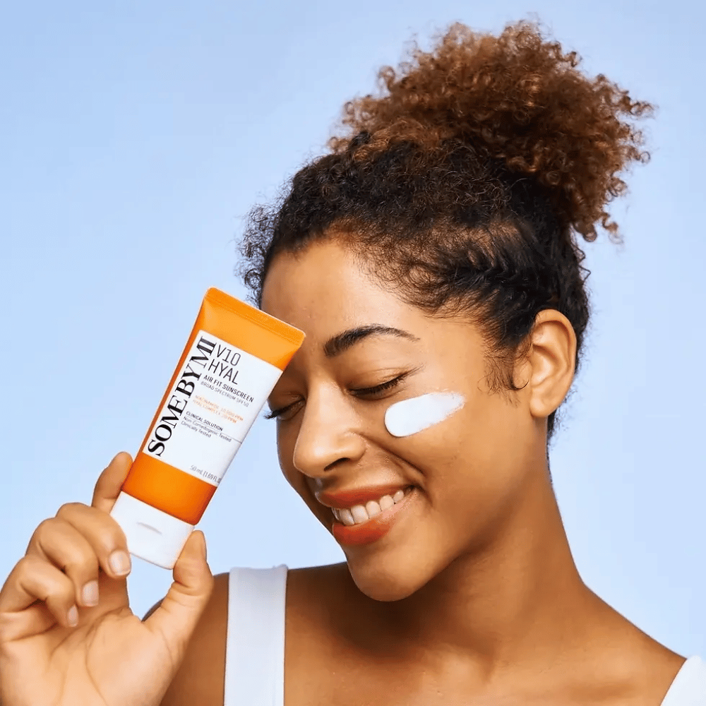 Ženska drži tubo SOME BY MI V10 Hyal Air Fit Sunscreen in ima belo liso kreme za zaščito pred soncem na licu. Veselo se smeji in jasno kaže izdelek, kar poudarja njegovo uporabo in prednosti za nego kože ter zaščito pred soncem.