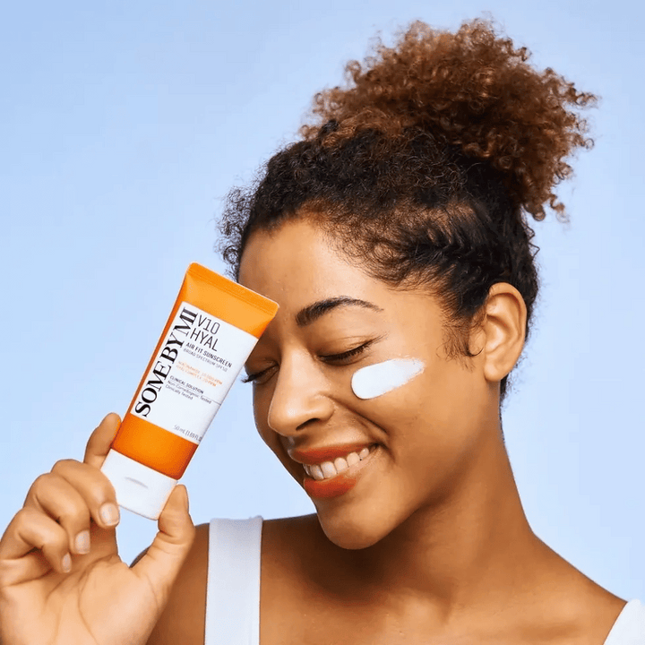 Ženska drži tubo SOME BY MI V10 Hyal Air Fit Sunscreen in ima belo liso kreme za zaščito pred soncem na licu. Veselo se smeji in jasno kaže izdelek, kar poudarja njegovo uporabo in prednosti za nego kože ter zaščito pred soncem.