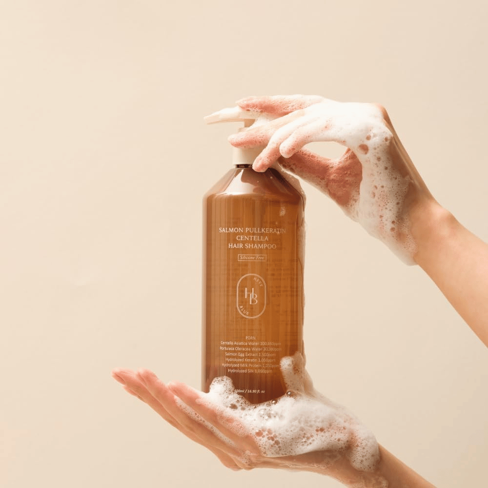 Heveblue Salmon Pullkeratin Centella Hair Shampoo v rjavi plastenki, obdani s peno. Izdelek je v rokah med dvema dlanema in poudarja čistilno ter negovalno izkušnjo nege las z obilico pene.