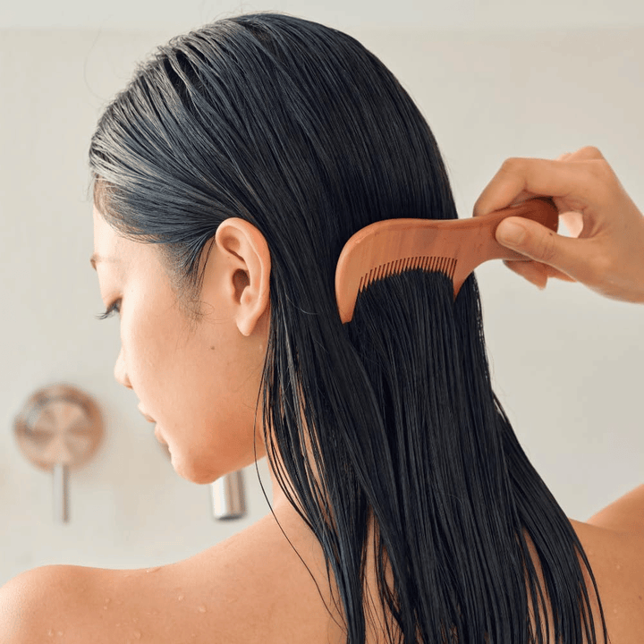 Oseba prečeše dolge, mokre lase s širokim glavnikom po nanosu Heveblue Salmon Pullkeratin Centella Hair Treatment. Prikazuje razčesavalni in mehčalni učinek za bolj gladek rezultat.