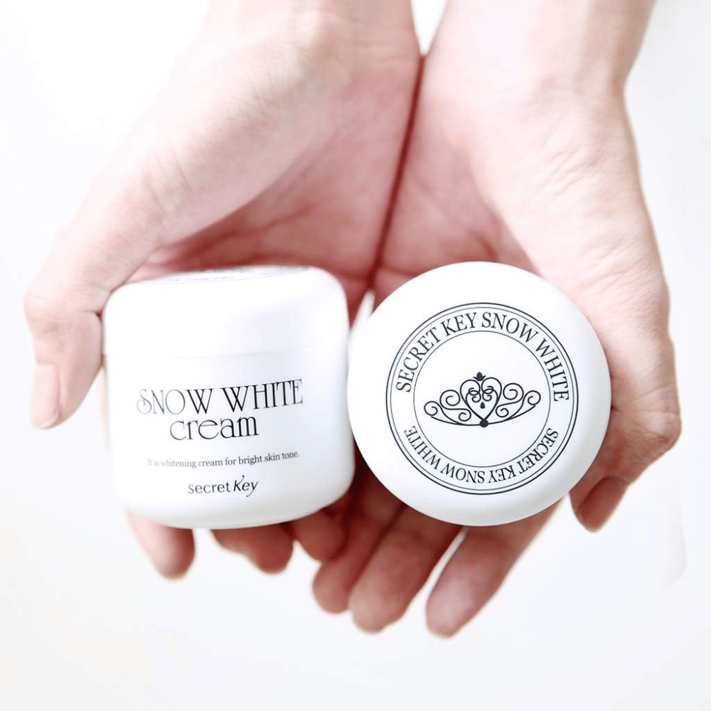Dve posodi Secret Key Snow White Cream držani v rokah, ena prikazuje pokrov in ena sprednjo stran.