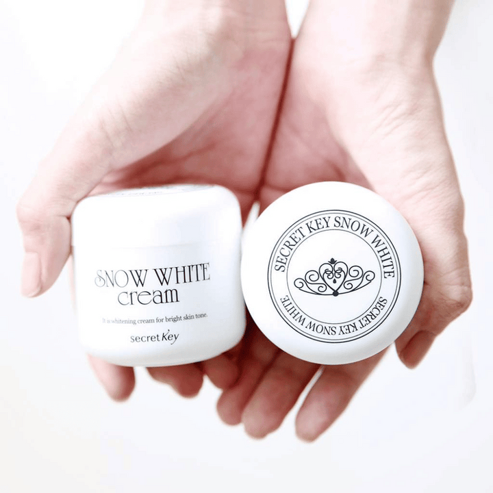 Dve posodi Secret Key Snow White Cream držani v rokah, ena prikazuje pokrov in ena sprednjo stran.