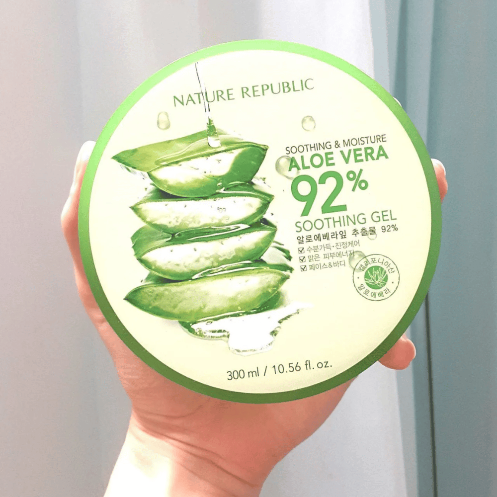 Roka drži okroglo posodo z Nature Republic Aloe Vera 92% Soothing Gel. Posoda je zelena s sliko aloe vere in besedilom, ki opisuje njene pomirjujoče in vlažilne lastnosti. Vsebina je 300 ml.
