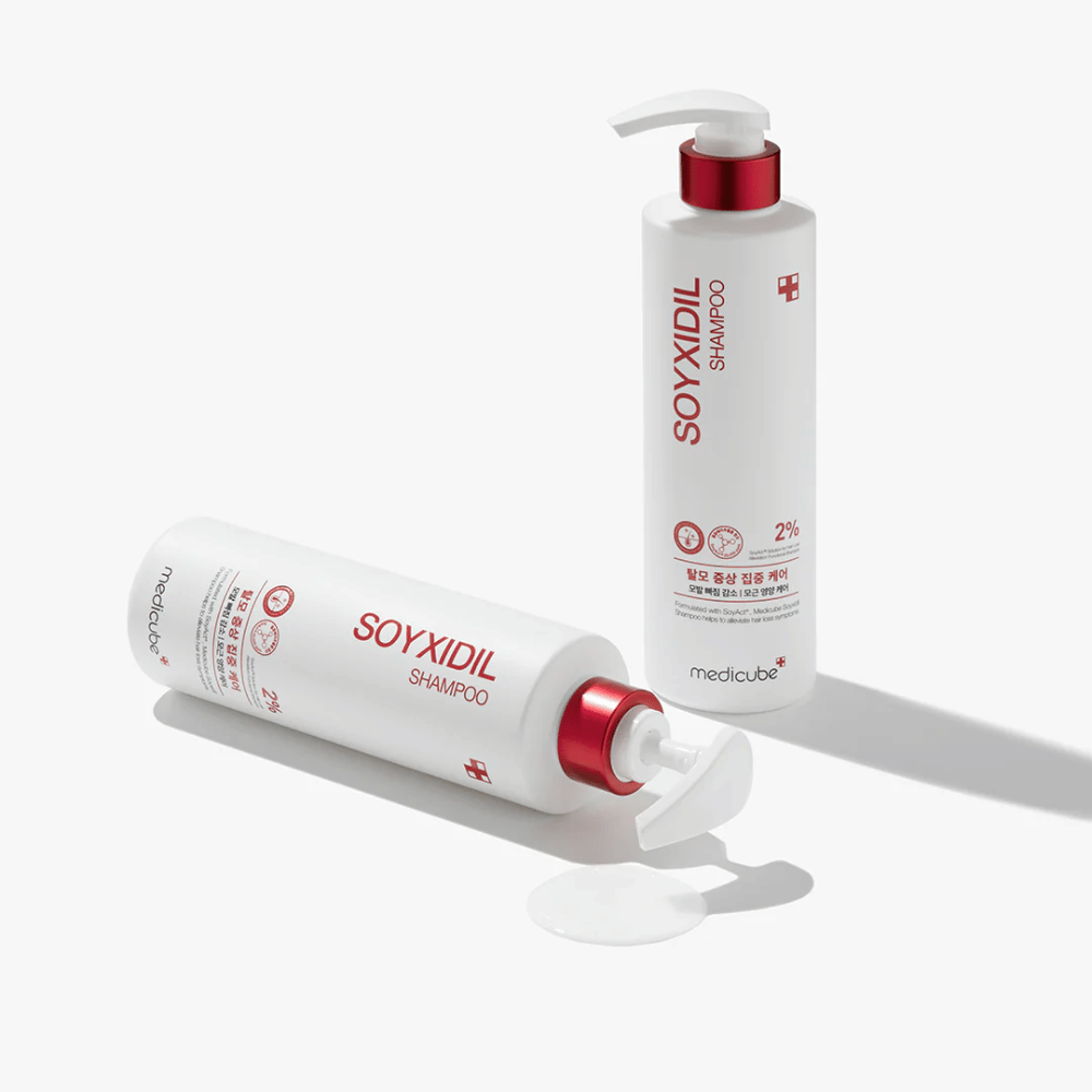 Dve steklenici Medicube Soyxidil Shampoo v beli in rdeči obliki, ena stoječa in ena ležeča s polito šamponom poleg.