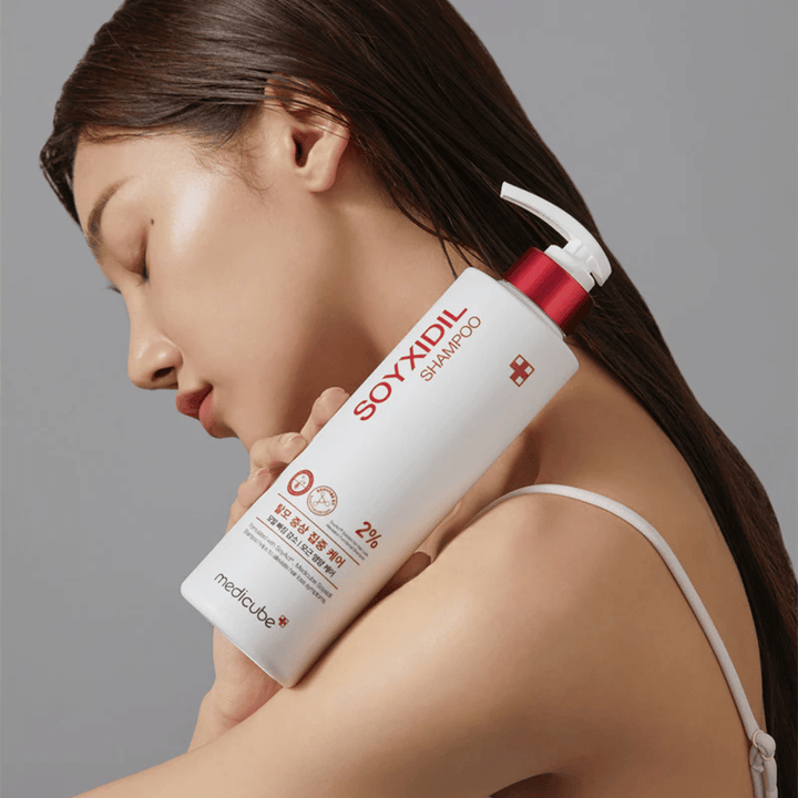 Ženska z dolgimi lasmi nagiba glavo na steklenico Medicube Soyxidil Shampoo, ki krepi lasišče in preprečuje izpadanje las.