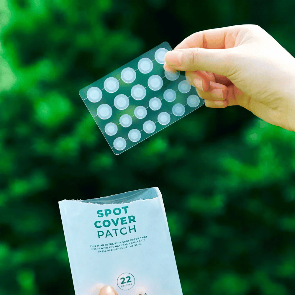 Roka drži list s Spot Cover obliži, ki so prozorni in zaobljeni. V ozadju je zamegljeno zeleno okolje. Embalaža prikazuje del embalaže z besedilom "SPOT COVER PATCH" in informacije, da izdelek vsebuje 22 obližev, ki pomagajo pri celjenju manjših kožnih površin.