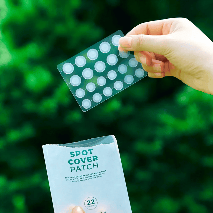Roka drži list s Spot Cover obliži, ki so prozorni in zaobljeni. V ozadju je zamegljeno zeleno okolje. Embalaža prikazuje del embalaže z besedilom "SPOT COVER PATCH" in informacije, da izdelek vsebuje 22 obližev, ki pomagajo pri celjenju manjših kožnih površin.