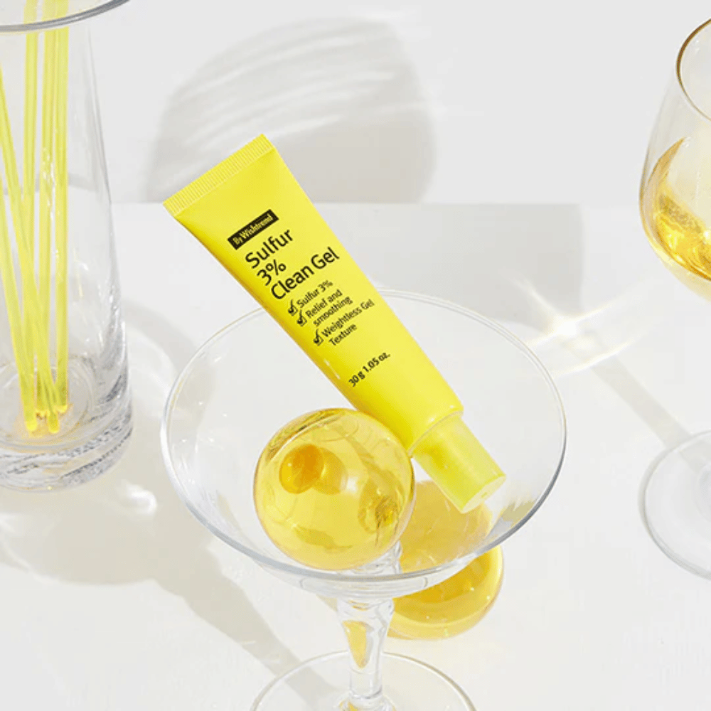 By Wishtrend Sulfur 3% Clean Gel v rumeni tubi, postavljeni v koktajl kozarec z rumeno sfero, obkroženo z elegantnimi kozarci na beli podlagi.