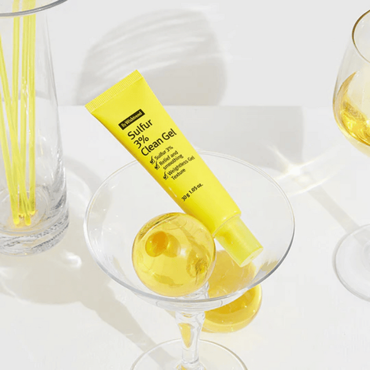 By Wishtrend Sulfur 3% Clean Gel v rumeni tubi, postavljeni v koktajl kozarec z rumeno sfero, obkroženo z elegantnimi kozarci na beli podlagi.