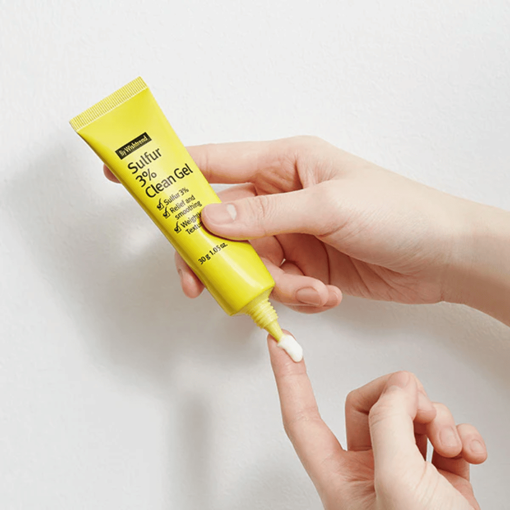 Bližnji posnetek rok, ki nanašajo By Wishtrend Sulfur 3% Clean Gel, rumena tuba z belo gelom, na beli podlagi.