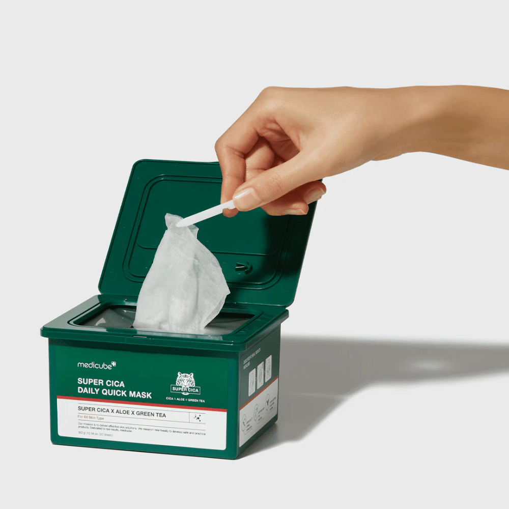 Zelena posoda z odprtim pokrovom prikazuje medicube Super Cica Daily Quick Mask. Roka izvleče masko iz tkanine s priloženimi pincetami. Izdelek vsebuje Cico, alojo in zeleni čaj, idealen za hitro vlaženje in pomirjujoč učinek.