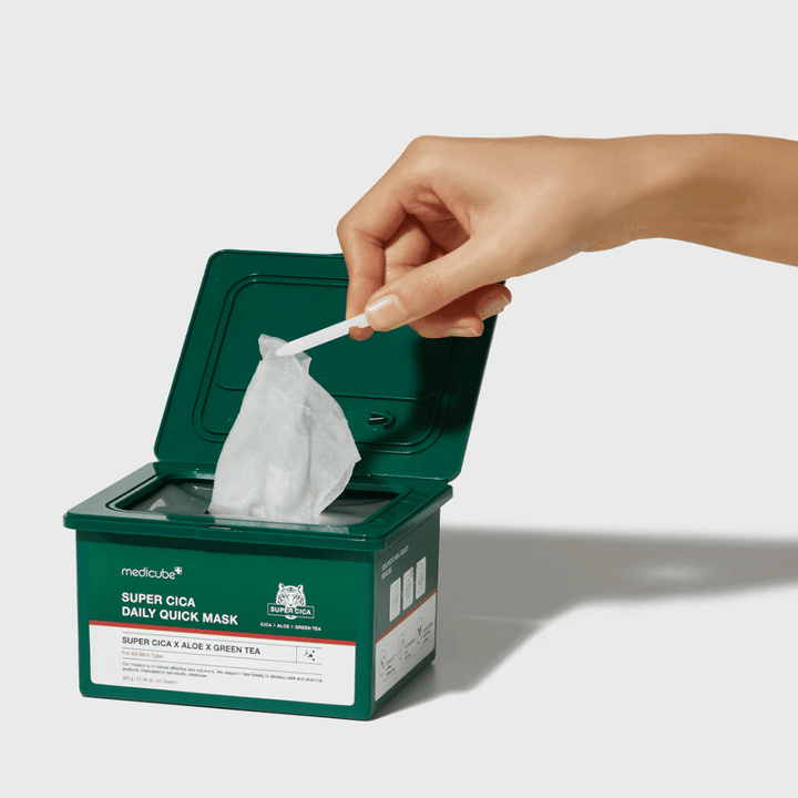 Zelena posoda z odprtim pokrovom prikazuje medicube Super Cica Daily Quick Mask. Roka izvleče masko iz tkanine s priloženimi pincetami. Izdelek vsebuje Cico, alojo in zeleni čaj, idealen za hitro vlaženje in pomirjujoč učinek.