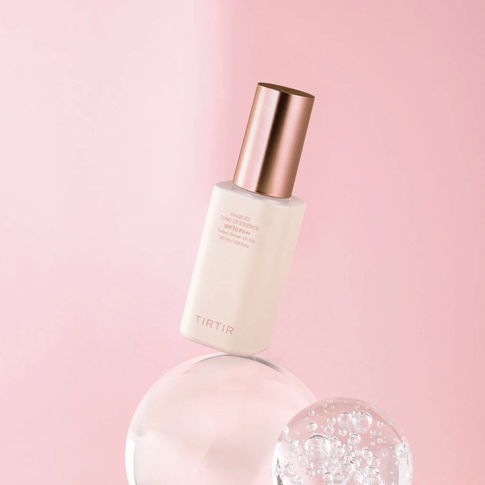Slika prikazuje stekleničko "TIRTIR MASK FIT TONE UP ESSENCE" z SPF30 PA++ na roza ozadju, kar daje občutek mehkobe in ženskosti.