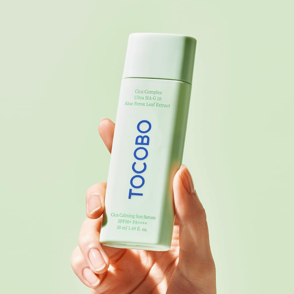 Roka drži TOCOBO Cica Calming Sun Serum z mintzeleno embalažo in modrim besedilom proti svetlozeleni podlagi.