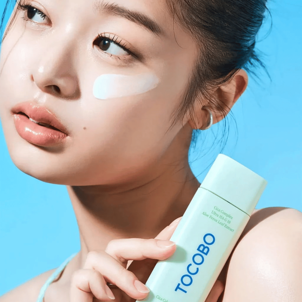 Model s sončno kremo pod očesom drži TOCOBO Cica Calming Sun Serum v mintzeleni embalaži proti modri podlagi.