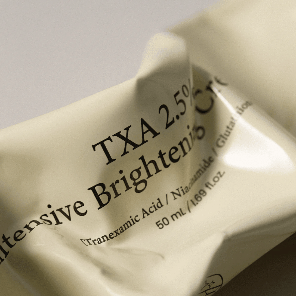 Bližinski posnetek AXIS - Y TXA 2.5% Intensive Brightening Cream, kjer je v ospredju mehak in minimalističen dizajn tube. Slika poudarja ime izdelka, Tranexamic Acid in Niacinamide ter elegantno bež embalažo.