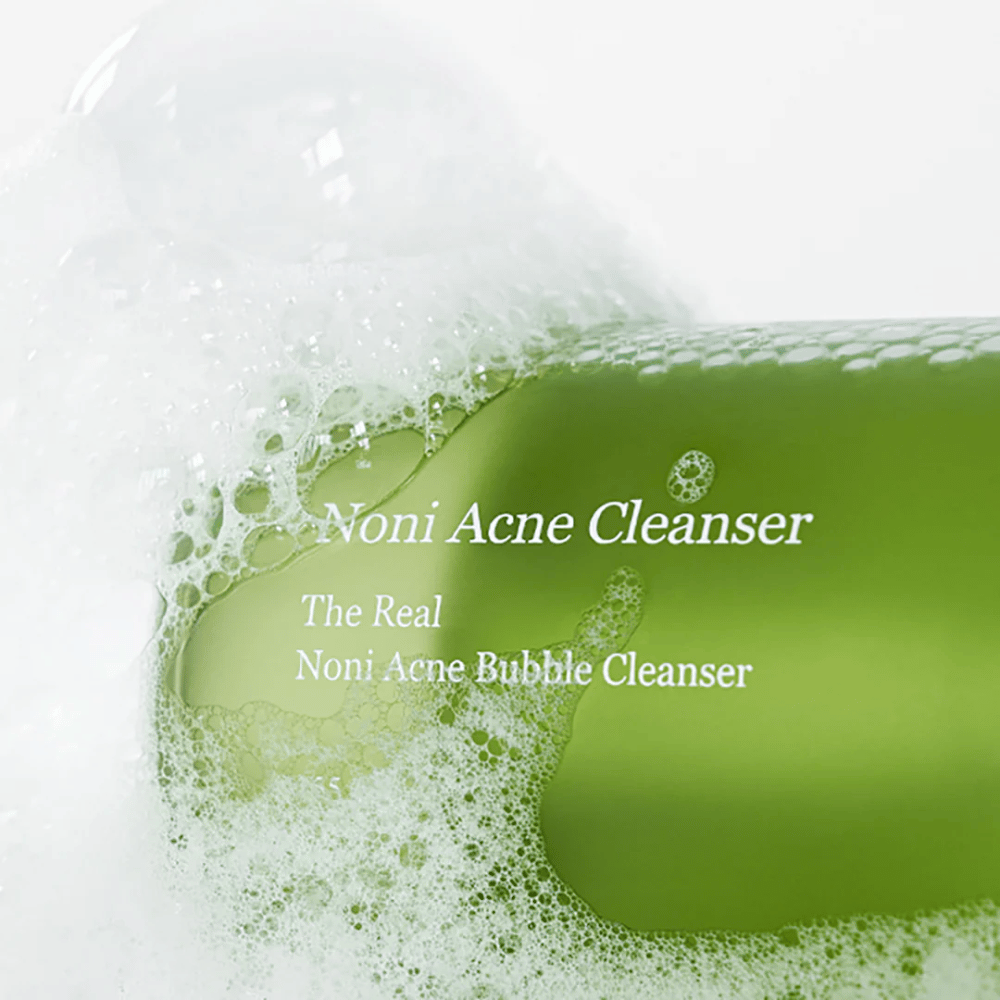Steklenica The Real Noni Acne Bubble Cleanser je v ospredju in obdana z obilico pene. Sveža zelena barva nakazuje naravne sestavine, popolne za čiščenje in boj proti aknam. Idealni izdelek za čisto in jasno kožo.