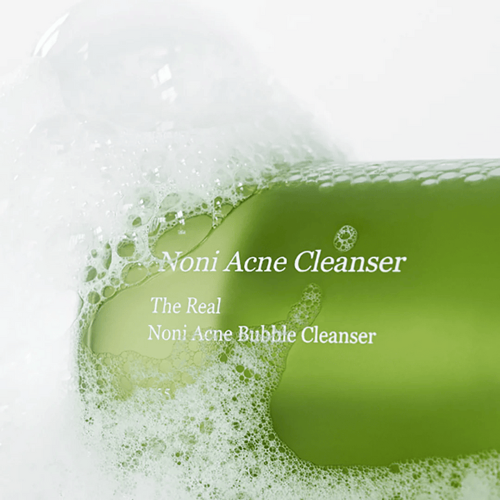 Steklenica The Real Noni Acne Bubble Cleanser je v ospredju in obdana z obilico pene. Sveža zelena barva nakazuje naravne sestavine, popolne za čiščenje in boj proti aknam. Idealni izdelek za čisto in jasno kožo.