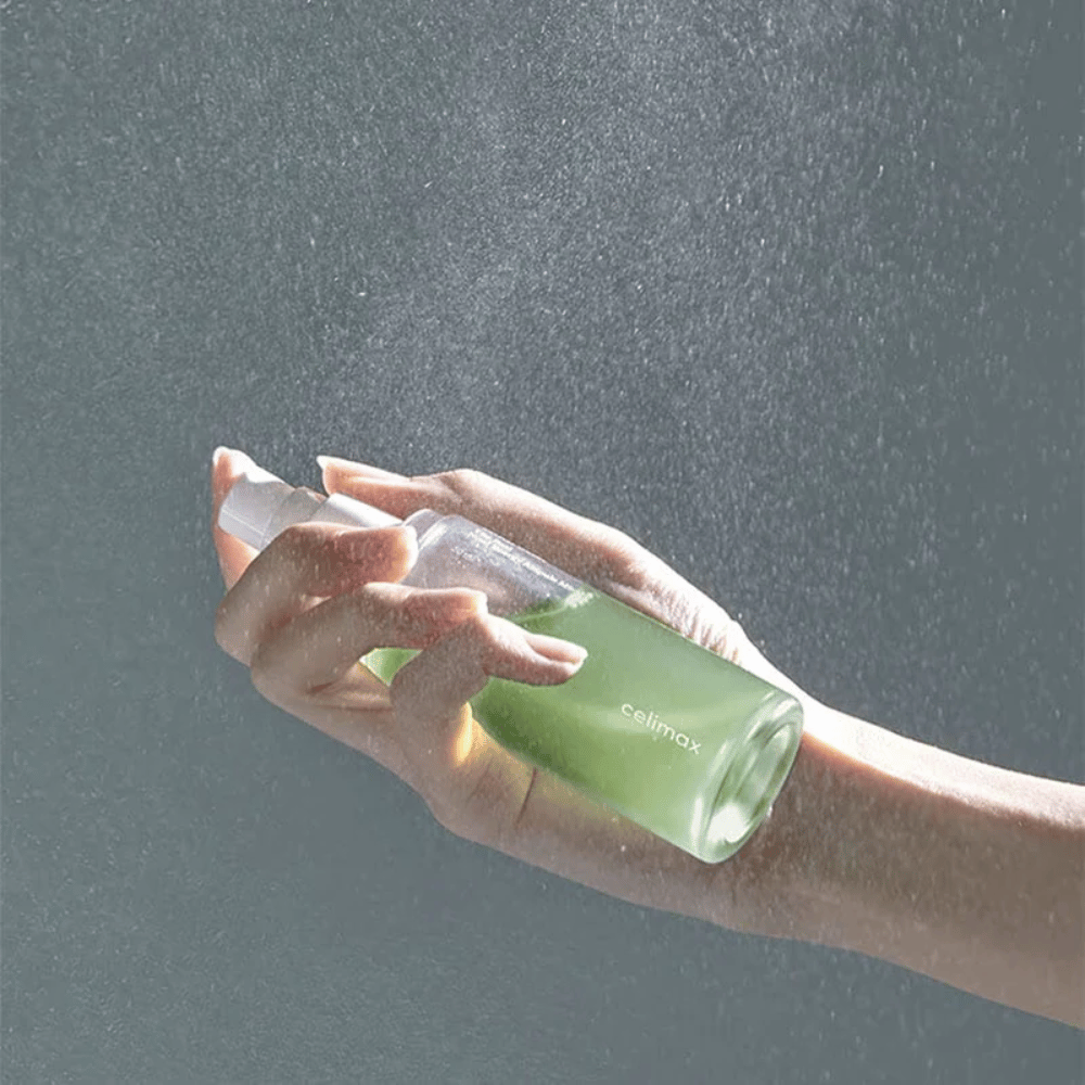 Roka drži razpršilno steklenico Celimax Noni Mist in nanaša izdelek v finem, enakomernem razpršilu na kožo. Steklenica je zelena in izdelek se trži kot "The Real Noni Energy Ampule Mist" za zagotavljanje energije in vlaženja kože.