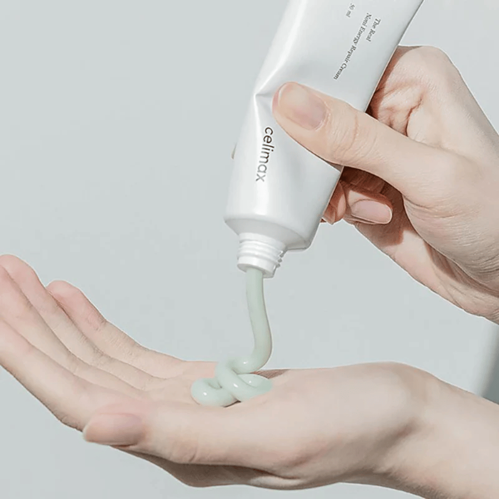Roka drži belo tubo s Celimax Noni Energy Repair Cream, krema pa se iztisne v enakomernem curku na dlan. Krema je svetlo zelena in je namenjena negi in obnovi kože.