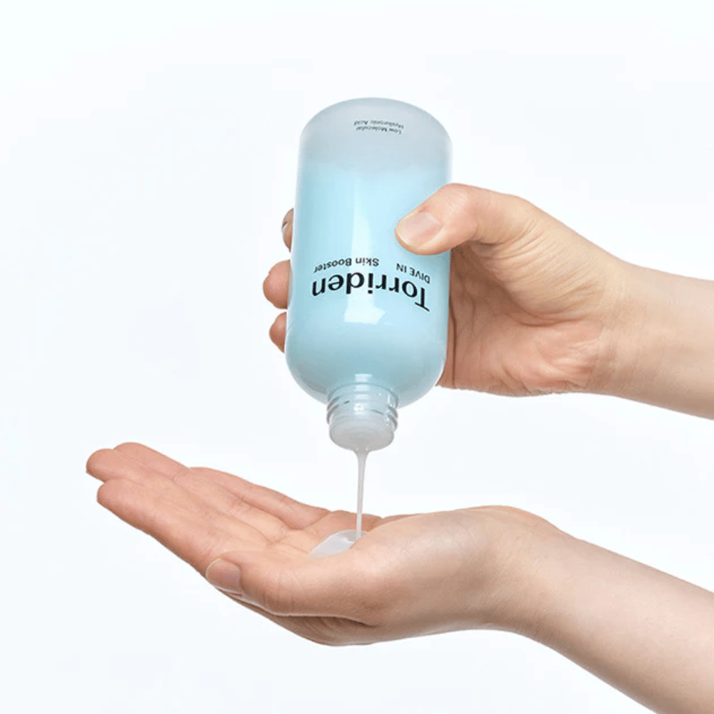 Roke, ki držijo steklenico s kozmetičnim izdelkom in dozirajo prozorno tekočino v dlan. Steklenica je označena z "Torriden - DIVE-IN Low Molecule Hyaluronic Acid Skin Booster".