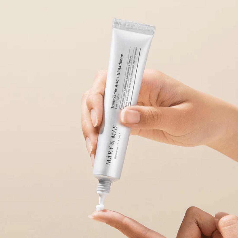 Roka drži tubo MARY & MAY "Tranexamic Acid + Glutathione Eye Cream". Tuba je bela, dizajn pa je čist in preprost, kar poudarja pomirjujoče in posvetlitvene lastnosti izdelka za področje okoli oči. Izdelek se nežno nanaša, s poudarkom na natančnosti in učinkovitosti.