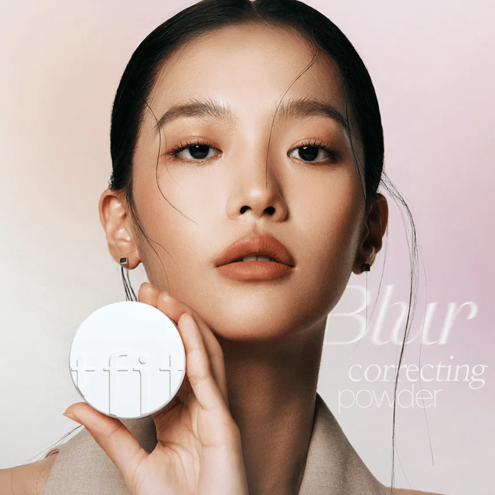 Ženska s popolnim ličenjem drži okroglo belo škatlico z logotipom TFIT, z napisom "Blur Correcting Powder" v ozadju.