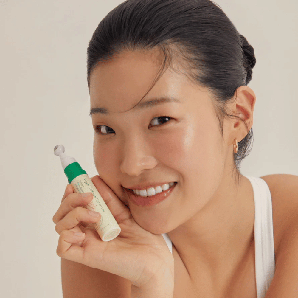 Ženska z dvignjenimi lasmi in zlatimi uhani drži AXIS-Y Vegan Collagen Eye Serum 10ml blizu obraza, medtem ko se nežno smeji. Serum je zasnovan za vlaženje in zmanjšanje gub okoli oči, posebej razvit za občutljivo kožo.