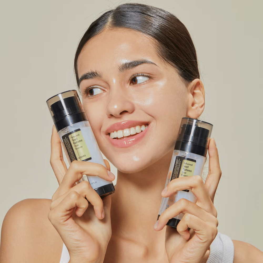 Nasmejana ženska z bleščečo kožo drži dve steklenici COSRX Advanced Snail Radiance Dual Essence 80 ml blizu obraza na svetlem ozadju.
