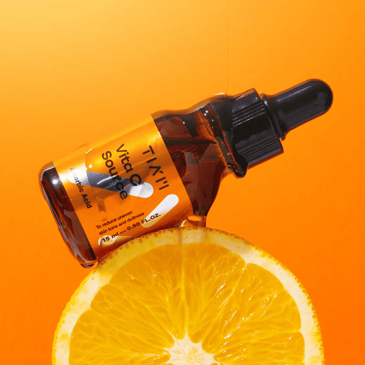 Steklenička TIA'M Vita C Source, postavljena ob pomarančo in oranžno ozadje, ki simbolizira svežino in antioksidativni učinek vitamina C.