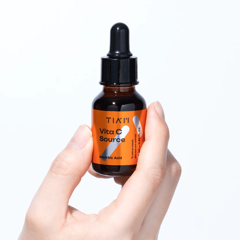 Roka drži TIA'M Vita C Source, serum z vitaminom C v rjavi steklenički s pipeto. Izdelek je zasnovan za izenačevanje tena kože in dodajanje sijaja.